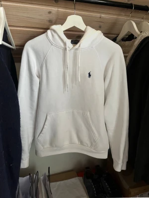 Vit hoodie från Ralph Lauren - Klassisk vit hoodie från Polo Ralph Lauren med blå logga broderad på bröstet. Hoodien har huva med dragsko, känguruficka och ribbade muddar. Tillverkad i mjukt bomullsmaterial som känns skönt mot huden. Perfekt för en clean och stilren look.