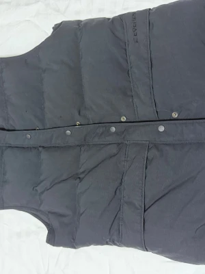 Mörkgrå dunväst från Everest - Snygg svart dunväst från Everest med GoldFlexSystem. Västen har tryckknappar framtill och två stora fickor. Perfekt för lager-på-lager och håller dig varm under kyliga dagar. Enkel, stilren design som funkar till det mesta.