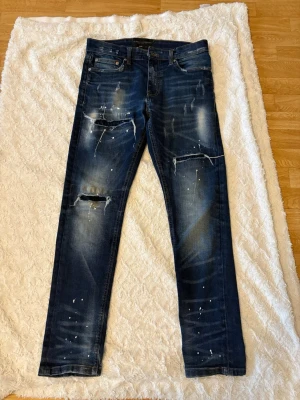 Mörkblå distressed jeans från Supply & Demand - Säljer ett par mörkblå jeans från Supply & Demand med slim fit passform. Jeansen har slitningar, hål vid knäna och vita färgstänk för en trendig, sliten look. Klassisk femficksmodell med dragkedja och knapp. Materialet är denim i bomull.