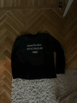 Svart långärmad Acne Studios t-shirt - Svart långärmad t-shirt från Acne Studios med trycket 'Acne Studios STOCKHOLM 1996' i vitt på bröstet. Klassisk rund hals och enkel design som passar till det mesta. Perfekt för dig som gillar stilrena plagg med en touch av streetwear.