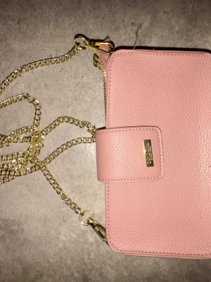 Rosa handväska från Don Donna  - Säljer en snygg ljusrosa handväska från Don Donna i strukturerat läder. Väskan har guldfärgade metalldetaljer och en kedjerem som ger en trendig look. Perfekt storlek för det viktigaste och stängs med magnetlås framtill.