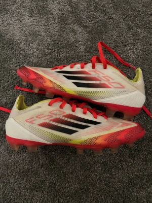 Adidas X F50 fotbollsskor - Säljer ett par Adidas X Speedflow fotbollsskor med snygg design i vitt, rött och gult. Skorna har svarta detaljer och röda snören samt dobbar för spel på gräs. Ovandelen är i lätt syntetmaterial för bra bollkontroll och snabbhet på planen.