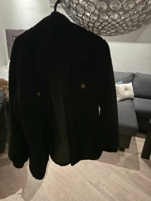 Svart teddyjacka med fickor - Mysig svart teddyjacka med två bröstfickor och knappar framtill. Jackan har en oversized passform och är gjord i fluffigt teddy-material som håller dig varm och stylish. Perfekt för att lyfta vilken outfit som helst under kyliga dagar.