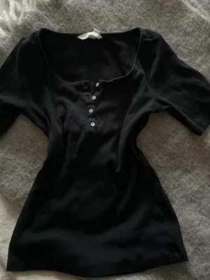 Bella swan/ elena Gilbert Svart ribbad topp med knappar  - Svart ribbad topp med djup rundad ringning och fyra vita knappar framtill. Toppen har korta ärmar och en figurnära passform som framhäver siluetten. Perfekt att styla med jeans eller kjol för en clean look.