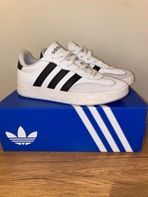 Adidas BARREDA SHOES - Snygga vita Adidas sneakers med klassiska svarta ränder på sidorna. Skorna har en låg silhuett, vita snören och detaljer i mocka framtill. Ovandelen är i läder och textil, och sulan är vit. Perfekta för en clean och sportig look. Använda ca 10 gånger. Storlek 42.