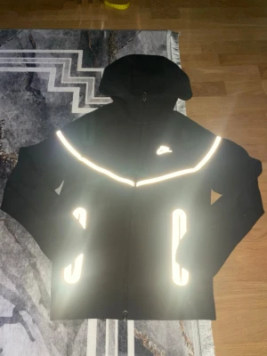 Svart Nike hoodie med reflexdetaljer - Snygg svart hoodie från Nike med dragkedja och huva. Hoodien har tydliga reflexdetaljer över bröstet och vid fickorna, samt en vit Nike-logga på vänstra sidan. Perfekt för dig som vill sticka ut och synas i mörkret. Tillverkad i mjukt material för extra komfort.