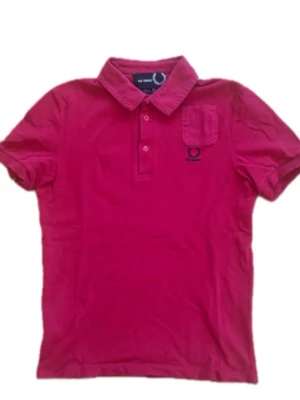 Röd pikéskjorta Raf Simons x Fred Perry - Sällsynt röd pikéskjorta från Raf Simons x Fred Perry med klassisk krage, kort ärm och bröstficka. Tillverkad i bomull och har knappar framtill. Perfekt för en clean och stilren look. OBS. Tveka inte på att höra av er vid frågor! Tröjan är i storlek 36 men skulle säga att den är S.