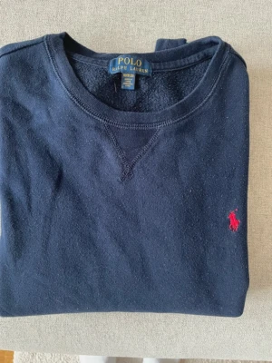Mörkblå sweatshirt från Polo Ralph Lauren - Klassisk mörkblå sweatshirt från Polo Ralph Lauren med rund halsringning och röd broderad logga på bröstet. Tröjan har långa ärmar och ribbade muddar vid ärmslut och nederkant. Perfekt för en avslappnad och stilren look.