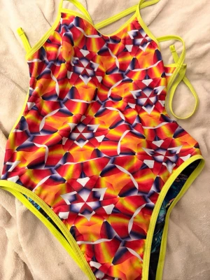 Speedo flipturn vändbar baddräkt  - Säljer en vändbar baddräkt med tunna axelband och två olika mönster: ena sidan har ett färgstarkt geometriskt mönster i rött, gult och blått, andra sidan har ett mörkt mönster med färgglada detaljer. Kanter och band är neongula. Perfekt för dig som vill sticka ut på stranden! Stl 28