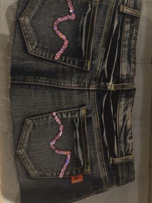 Jeanskjol med paljettdetaljer - Säljer ett par mörkblå jeansshorts med slitna kanter och coola paljettdetaljer i rosa och grönt på fickorna. Shortsen har klassisk femficksdesign och kontrastsömmar. Perfekta för dig som vill sticka ut lite extra i sommar!