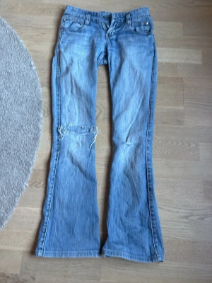 Ljusblå bootcut jeans med hål - Säljer ett par ljusblå bootcut jeans med slitning och hål på ena knät. Jeansen har klassisk femficksmodell och är gjorda i denim med snygg tvätt. Perfekta för en avslappnad och trendig look. Bara att gylfen är trasig därför dras priset ned.!