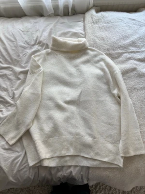 Vit polotröja i stickat material - Klassisk vit stickad polotröja med bred ribbad krage, muddar och nederkant. Tröjan har en avslappnad passform och är perfekt för kyliga dagar. Materialet ser mjukt och bekvämt ut, och färgen är vit. Sitter mer som M