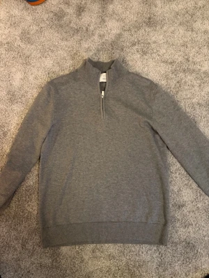 Grå half zip tröja från Bläck - Snygg grå tröja från Bläck med halv dragkedja och hög krage. Tröjan har ribbade muddar vid ärmslut och nederkant, perfekt för en clean och stilren look. Tillverkad i mjukt material som känns skönt mot huden.