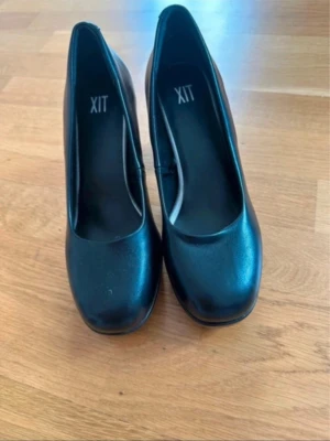 Svarta pumps med klack från XIT - Stilrena svarta pumps från XIT med rundad tå och bred klack. Skorna har en klassisk design i slätt material som ger en tidlös look. Perfekta för dig som vill ha ett par enkla men snygga klackar till garderoben.