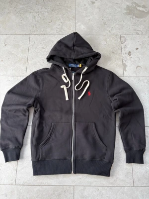 Svart hoodie från Polo Ralph Lauren - Säljer en svart hoodie från Polo Ralph Lauren med dragkedja, vit dragsko och klassisk röd logga på bröstet. Hoodien har känguruficka framtill och ribbade muddar vid ärmar och nederkant. Tillverkad i mjuk bomullsblandning för extra komfort.