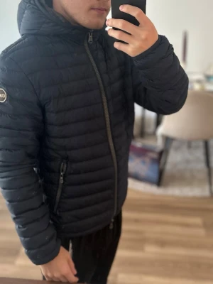 Mörkblå dunjacka med huva från Colmar - Säljer en svart dunjacka från Colmar med huva och dragkedja framtill. Jackan har två sidofickor med dragkedja och ett Colmar-märke på ärmen. Lätt vadderad och quiltad design som håller värmen under kalla dagar. Storlek 46.