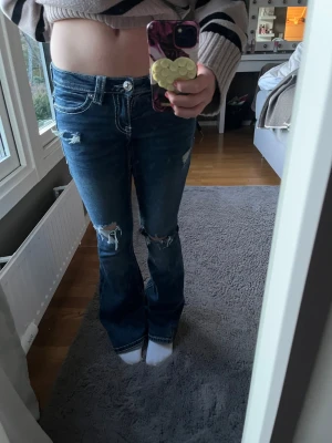 Blå bootcut jeans med slitningar - Säljer ett par blå bootcut jeans med slitningar på knäna och snygga kontrastsömmar. Jeansen har låg midja och dekorativa broderier på bakfickorna. Perfekta för en avslappnad och trendig look.