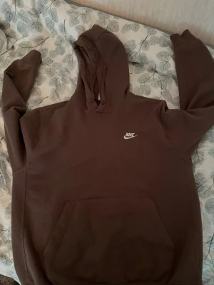 Brun hoodie från Nike med huva - Säljer en brun hoodie från Nike med klassisk vit logga på bröstet. Hoodien har en stor känguruficka framtill och justerbar huva med dragsko. Tillverkad i mjukt material som känns skönt mot huden. Perfekt för en avslappnad och sportig stil.