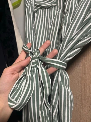 Grönvit randig skjortklänning med knyt - Snygg grön och vit randig skjortklänning med knappar framtill och knytband i midjan från vero Moda . Klänningen har krage och bröstfickor, vilket ger en avslappnad men ändå chic look. Perfekt för dig som gillar stilrena detaljer och mönster. Aldrig använt då den inte är min stil men super fin för sommaren 