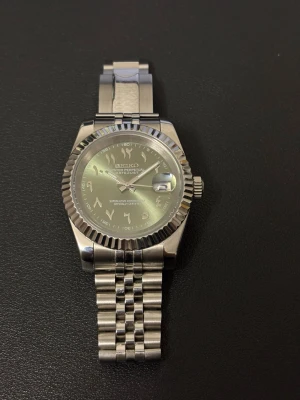 Seiko mod klocka - Stilren Seiko klocka i silver med klassisk länkarmband och räfflad bezel. Urtavlan har arabiska siffror och datumfönster med förstoringsglas. Klockan har en elegant och tidlös design som passar till många olika stilar.
