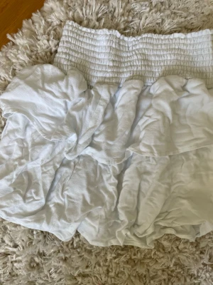 Vit volangkjol med smock - Söt vit kortkjol med bred smock i midjan och volangdetaljer. Kjolen är gjord i ett lätt och luftigt material, perfekt för varma dagar. Passar till både sneakers och sandaler för en avslappnad look.