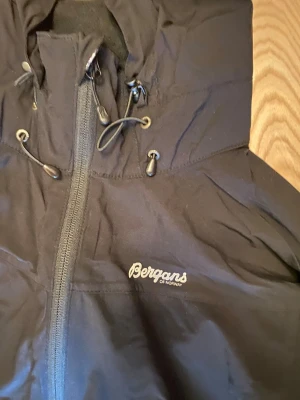 Svart vindjacka från Bergans - Snygg svart vindjacka från Bergans of Norway med huva och dragsko. Jackan har hel dragkedja framtill, två sidofickor med dragkedja och justerbara ärmslut. Perfekt för blåsiga dagar och outdoor-aktiviteter.