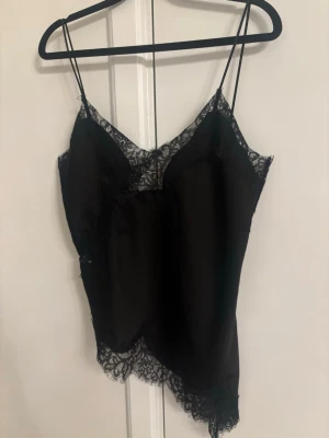 Svart spetsdetaljerad linnetopp Zara - Snygg svart linnetopp med tunna axelband och spetsdetaljer längs urringning och nederkant. Toppen har en asymmetrisk form och är tillverkad i ett glansigt material som ger en lyxig känsla. Perfekt för dig som vill ha en trendig och elegant look. Helt ny med prislapp slutsåld överallt viral Zara topp