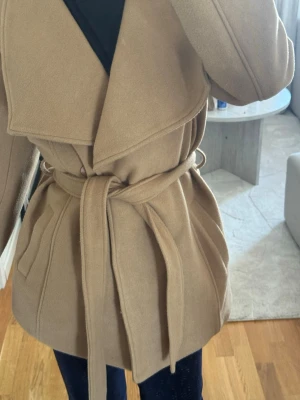 Beige trenchcoat med knytskärp - Stilren beige trenchcoat med bred krage och knytskärp i midjan. Jackan har två fickor framtill och är tillverkad i ett mjukt tyg som ger en klassisk look. Perfekt för dig som vill ha en tidlös och trendig jacka till hösten. Aldrig använd, köpt för 999kr