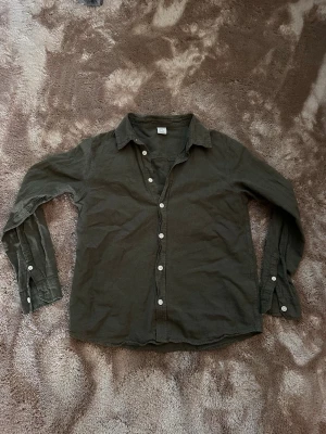 Olivgrön linneskjorta  - Snygg olivgrön skjorta i linne med klassisk krage och vita knappar hela vägen framtill. Skjortan har lång ärm och en avslappnad passform, perfekt för en chill och stilren look.