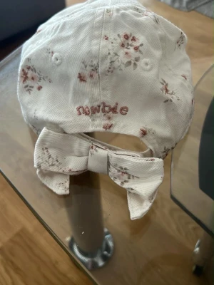 Blommig keps med rosett från Newbie - Söt keps från Newbie i ljus bomull med små blommönster i rosa och beige toner. Baktill finns en stor dekorativ rosett och broderad logga. Passar barn mellan 9 månader och 3 år. Perfekt accessoar för soliga dagar.