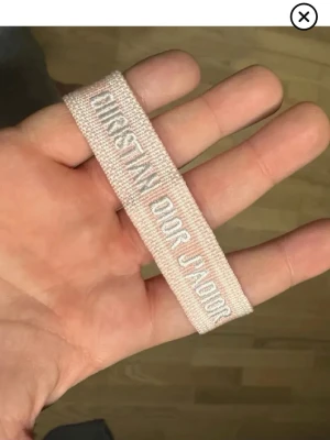 Christian Dior rosa tygarmband - Säljer ett ljusrosa tygarmband från Christian Dior med texten 'Christian Dior J'Adior' broderat i vitt. Armbandet har justerbara knytband och är tillverkat i mjukt vävt material. Perfekt accessoar för dig som gillar exklusiva detaljer och vill lyfta din outfit.