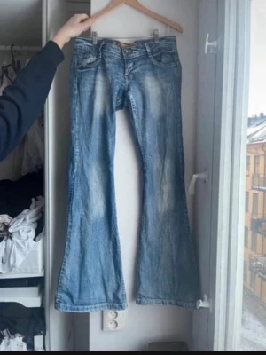 Blå bootcut jeans med slitningar - Säljer ett par blå bootcut jeans med snygga slitningar och ljusare partier framtill. Jeansen har låg midja, klassiska fem fickor och dekorativa sömmar på bakfickorna. Perfekta för dig som gillar en retro och avslappnad stil.