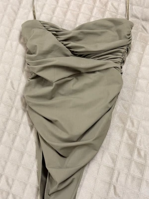 Armegrön axelbandslös body - Trendig armegrön axelbandslös body/topp med omlottdesign och draperade detaljer. Bodyn har en asymmetrisk form och är tillverkad i ett mjukt tyg som ger en snygg siluett. Perfekt för dig som vill sticka ut med en stilren look.