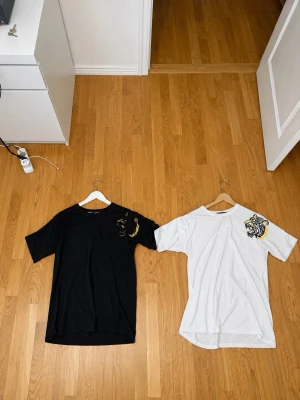 Philipp Plein t-shirts med tigertryck - Två snygga t-shirts från Philipp Plein, en svart och en vit, båda med coolt tigertryck på bröstet. Klassisk rund hals och korta ärmar. Perfekta för dig som vill sticka ut med streetwear-vibe. Tillverkade i mjuk bomull för skön känsla.
