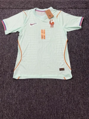 Ljusblå Frankrike Nike matchtröja - Snygg ljusblå matchtröja från Nike med Frankrikes landslagsemblem och nummer 11 i orange framtill. Tröjan har korta ärmar med randiga muddar i blått, vitt och rött samt orange detaljer längs sidorna. Tillverkad i lätt och ventilerande material, perfekt för fotboll.
