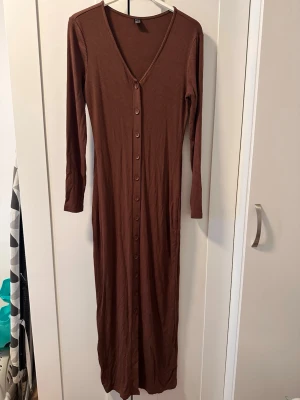 Brun ribbad långklänning med knappar - Säljer en brun långklänning med ribbad struktur och v-ringning. Klänningen har långa ärmar och knappar hela vägen framtill, vilket ger en snygg och enkel look. Perfekt för dig som gillar stilrena och bekväma plagg.