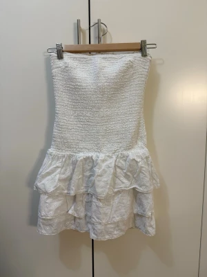 Vit axelbandslös volangklänning - Söt vit kort klänning med smockad överdel och volanger i kjolen. Axelbandslös modell med broderade detaljer på kjolen som ger en romantisk känsla. Perfekt för varma dagar och somriga tillfällen.