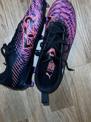 Puma Future Z Svart/Lila Fotbollsskor - Snygga fotbollsskor från Puma Future Z-serien i svart med lila och rosa mönster. Skorna har snörning, syntetiskt material och dobbar för spel på gräs. Perfekta för dig som vill sticka ut på planen med både stil och prestanda.