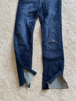 Mörkblå jeans från Denim Deluxe - Säljer ett par mörkblå jeans från Denim Deluxe med raka ben och slitningar på ena låret. Jeansen har råa, avklippta benslut och klassiska kontrastsömmar. Perfekta för dig som gillar en edgy och avslappnad stil.