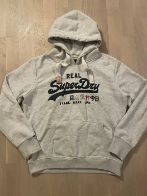 Superdry hoodie - Snygg grå hoodie med vita snören och snygga detaljer från SUPERDRY. Hoodien passar storlek S, men det står L i den. I mycket bra skick, iprincip oanvänd och inga defekter alls! Hör av er vid frågor!🔥🤝