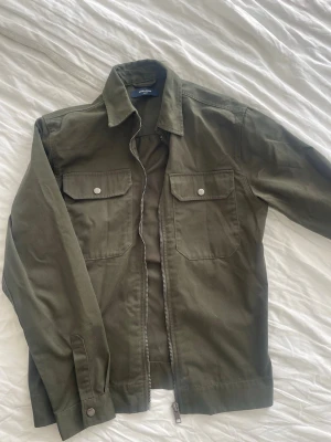 Olivgrön overshirt från jack and jones - Stilren olivgrön overshirt från jack and jones med två bröstfickor med lock och tryckknappar. Klassisk krage och dragkedja framtill. 