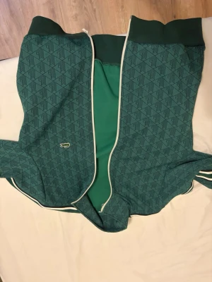 Grön mönstrad hoodie från Lacoste - Snygg grön hoodie från Lacoste med diskret mönster i mörkare nyans och vit piping längs dragkedjan. Klassisk krokodil-logga på bröstet och ribbade muddar. Tillverkad i mjukt bomullsmaterial som känns skönt mot huden. Perfekt för en chill och sportig look.