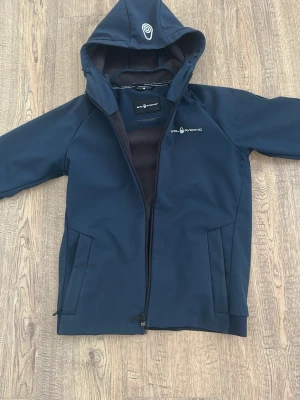 Mörkblå vindjacka från Sail Racing - Stilren mörkblå vindjacka från Sail Racing med huva och dragkedja framtill. Jackan har två sidofickor och mjukt fleecefoder på insidan för extra komfort. Perfekt för blåsiga dagar och har en sportig look med logga på bröstet och huvan.