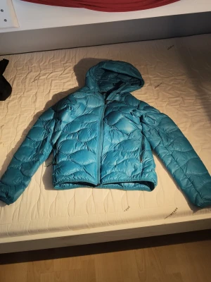 Blå dunjacka med huva Peak Performance - Snygg blå dunjacka från Peak Performance med huva och dragkedja framtill. Jackan har ett quiltat mönster och två sidofickor med dragkedja. Perfekt för kalla vinterdagar och riktigt lätt att bära. Klassisk modell som håller värmen bra.