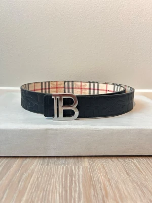 Svart Burberry bälte med B-spänne - Snyggt svart bälte från Burberry med ett stort silverfärgat B-spänne. Bältet har ett diskret mönster på utsidan och klassiskt Burberry-randigt foder på insidan. Perfekt accessoar för att lyfta din outfit.