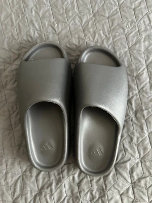 Yeezy Slides  - Säljer ett par stilrena gråa slides från Adidas. Tofflorna har en chunky sula med räfflad undersida och bred rem över foten. Tillverkade i mjukt och flexibelt syntetmaterial för maximal komfort. Perfekta för chill dagar hemma eller till stranden.