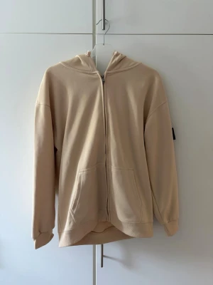 Beige zip hoodie från Stone Island - Snygg beige hoodie från Stone Island med dragkedja framtill och klassisk patch på ärmen. Tillverkad i mjuk bomull, har två fickor fram och ribbade muddar. Perfekt för dig som gillar streetwear och vill ha en clean look.