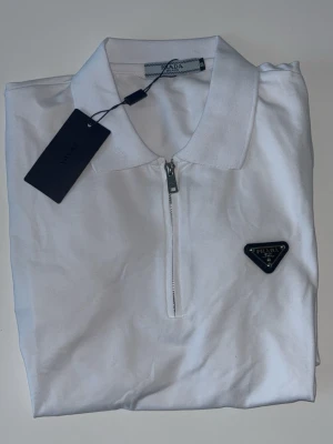 Vit half zip t-shirt från Prada - Snygg vit t-shirt från Prada med klassisk krage och half zip-dragkedja framtill. T-shirten har en diskret logga på bröstet och är tillverkad i mjuk bomull för en clean och stilren look.