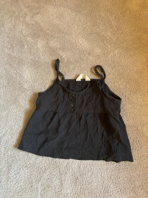 Svart ribbad crop top med knappar från H&M i strl 146 - Snygg svart crop top med tunna axelband och ribbad struktur. Toppen har fyra dekorativa knappar framtill och en avslappnad passform. Perfekt till jeans eller kjol för en trendig look.