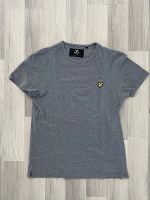 Grå t-shirt från Lyle & Scott - Snygg grå t-shirt från Lyle & Scott med klassisk rund hals och korta ärmar. T-shirten har den ikoniska gula loggan broderad på bröstet. Tillverkad i mjuk bomull för en bekväm känsla och enkel stil.